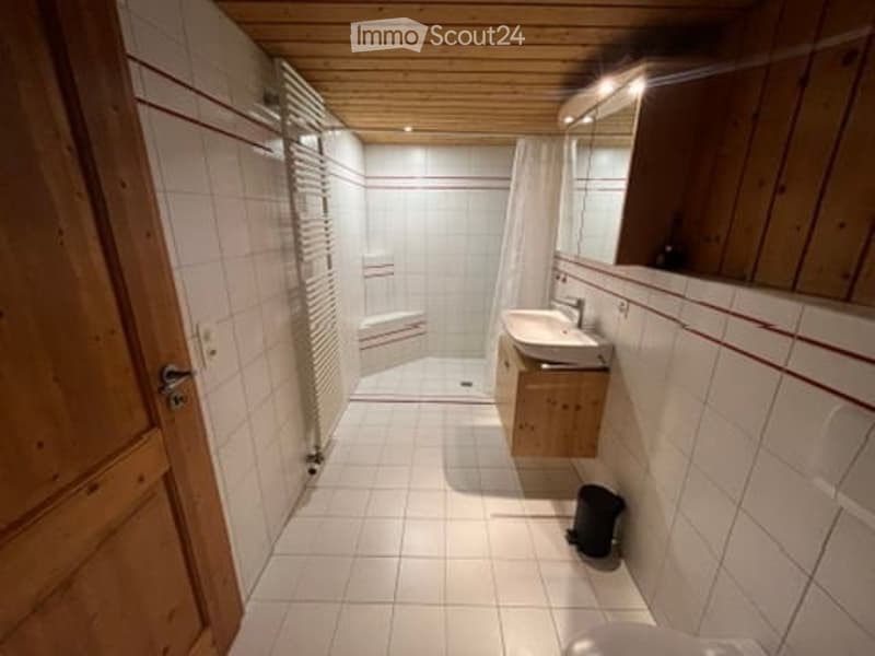 Modern bathroom with white tiles, wooden ceiling, and a large mirror above the sink. Features a shower area with a curtain and a small trash bin on the floor.