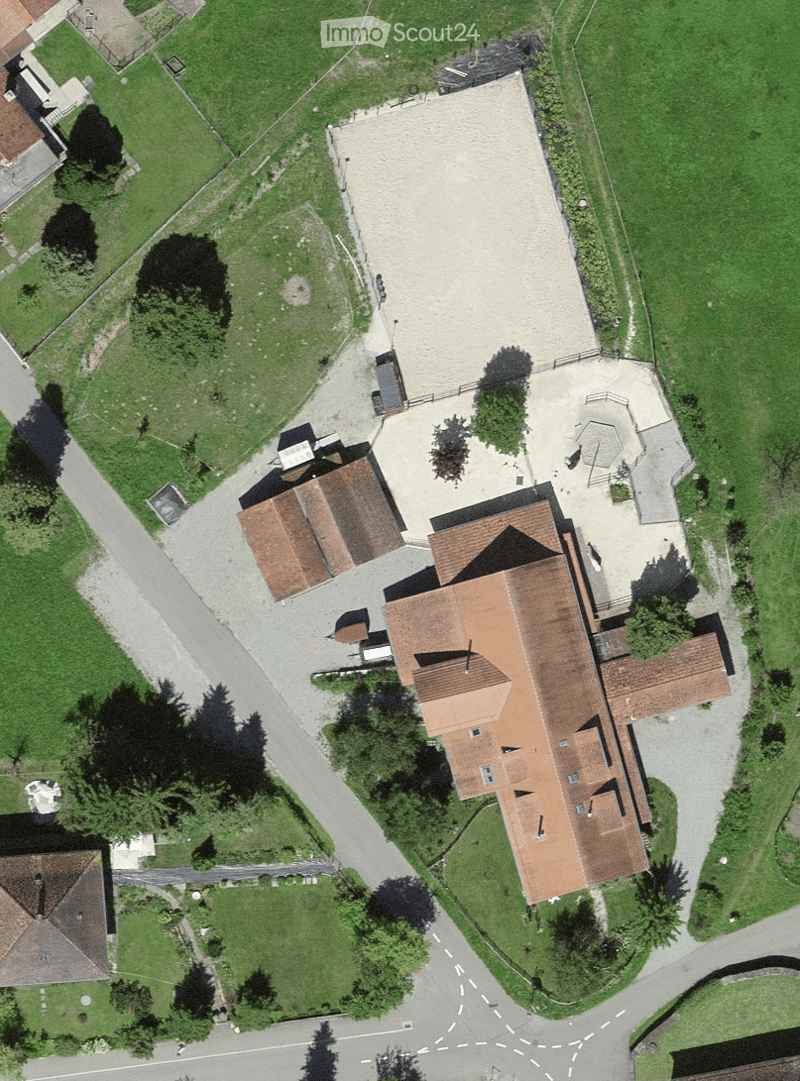 Aerial view of a large building complex with multiple structures, surrounded by green fields and trees. The main building has a red-tiled roof and is connected to smaller outbuildings and paved areas.