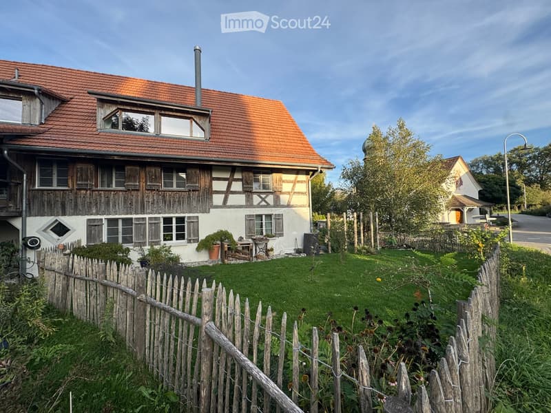 Traditional two-story house with a red-tiled roof, wooden facade, and multiple windows. The house is surrounded by a wooden fence and a well-maintained lawn.