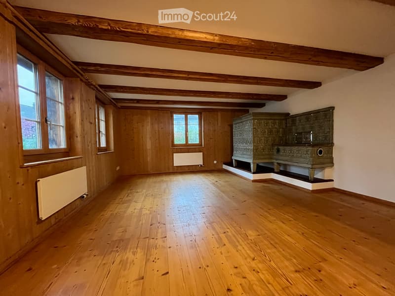 An empty room with wooden floors, exposed wooden beams, and large windows. The room features wooden panel walls and radiators along the walls.
