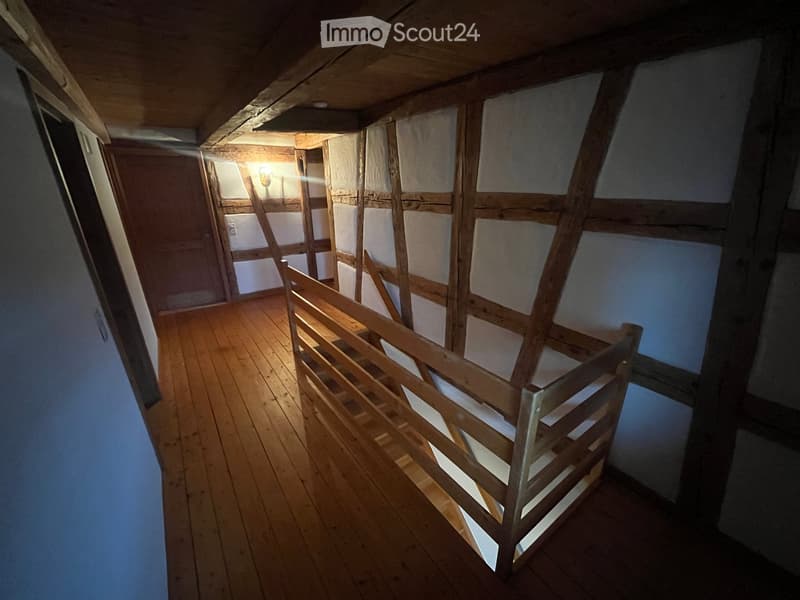 Wooden staircase with a simple wooden railing, leading up to an attic space with exposed wooden beams and unfinished walls.