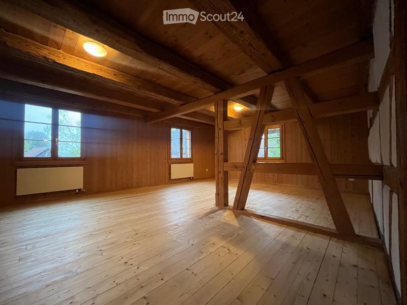 Empty attic space with wooden beams, wooden floor, and large windows allowing natural light. Exposed wooden beams and simple wooden walls.
