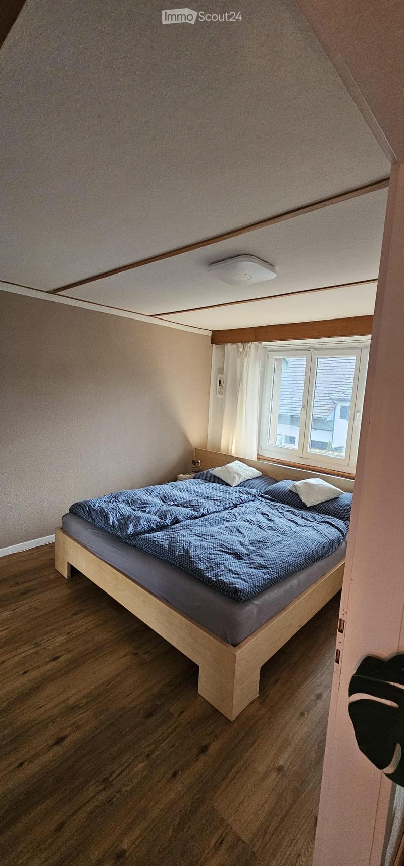 Cozy attic bedroom with wooden floor, slanted ceiling, and a single bed with blue bedding and white pillows. A window with white curtains offers natural light.