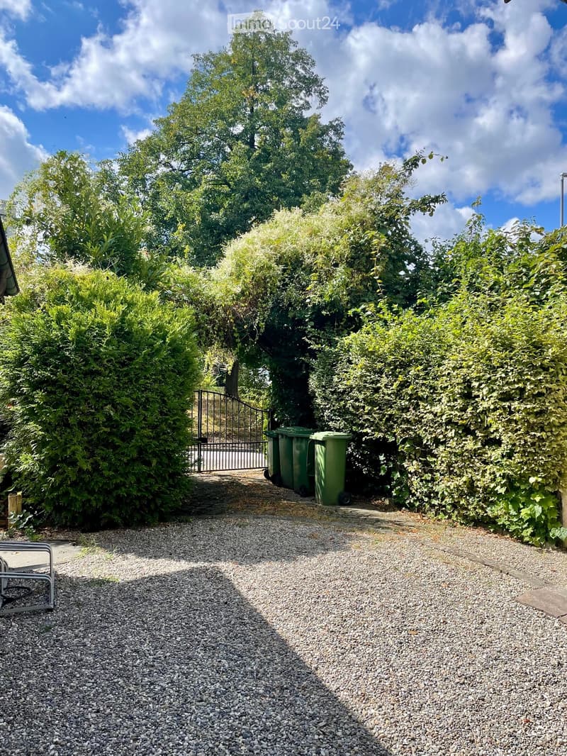 Gravel pathway leading to a gated entrance, surrounded by lush green bushes and trees, with green recycling bins placed near the gate.
