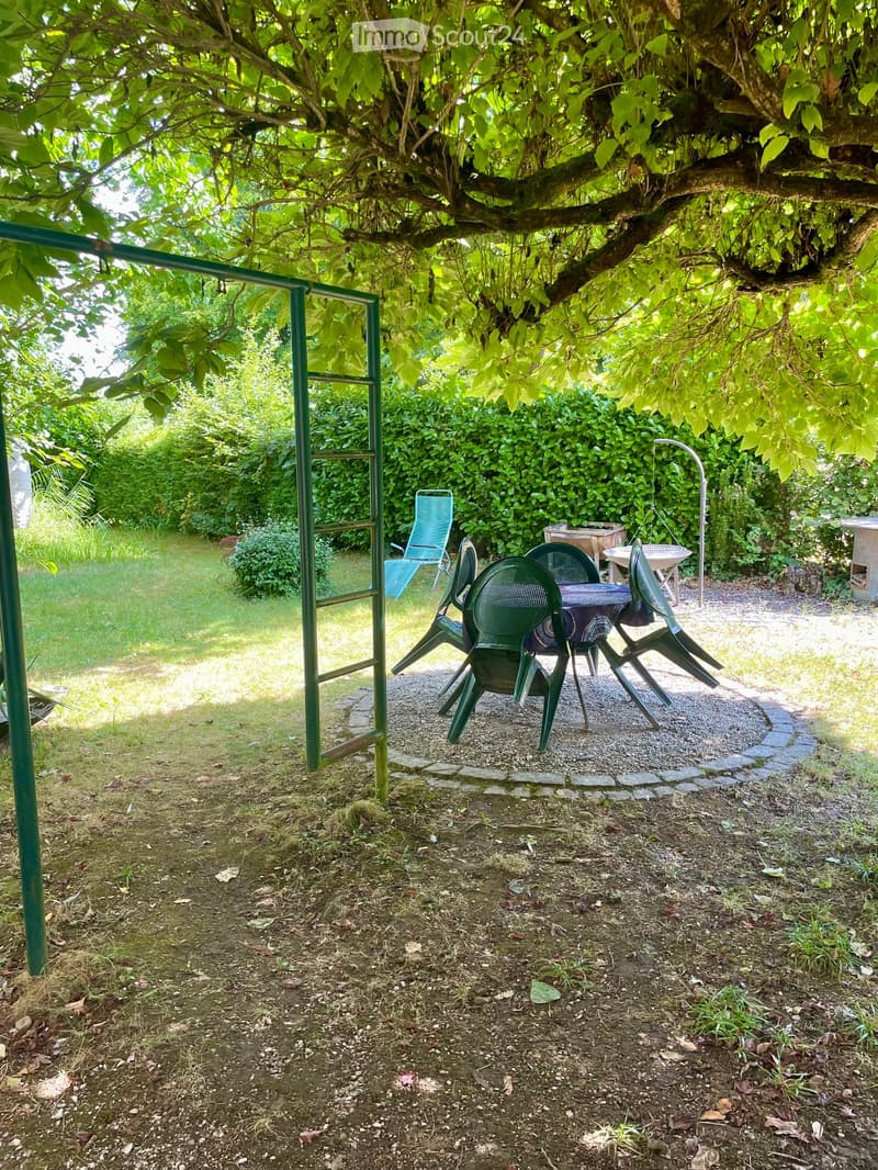 A shaded outdoor seating area with a table and chairs under a large tree, surrounded by greenery and a gravel path.