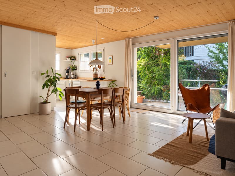 Bright, open-plan living and dining area with wooden dining table, chairs, sofa, pendant light, and large windows overlooking a garden.