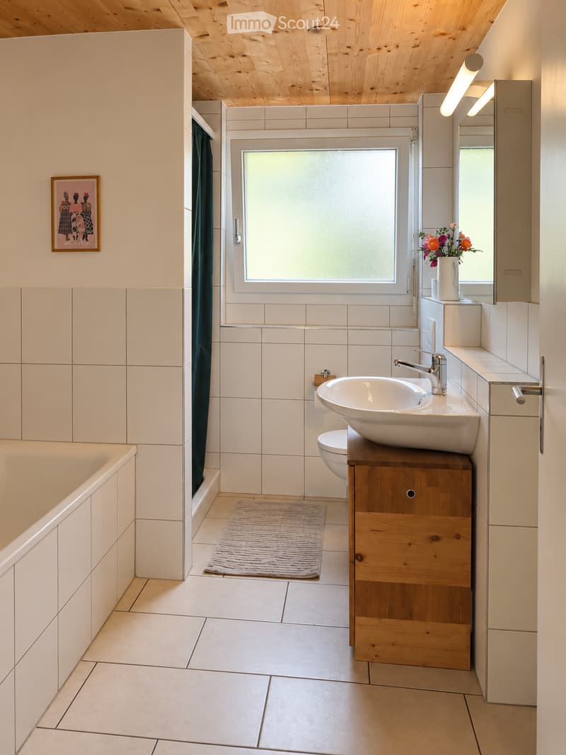 Modern bathroom with white tiles, wooden ceiling, bathtub, toilet, sink, mirror, and a small wooden cabinet.