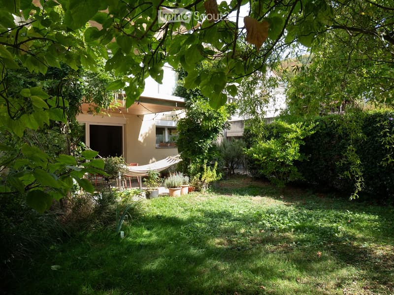 A lush garden with a hammock, potted plants, and a small seating area near the house entrance.