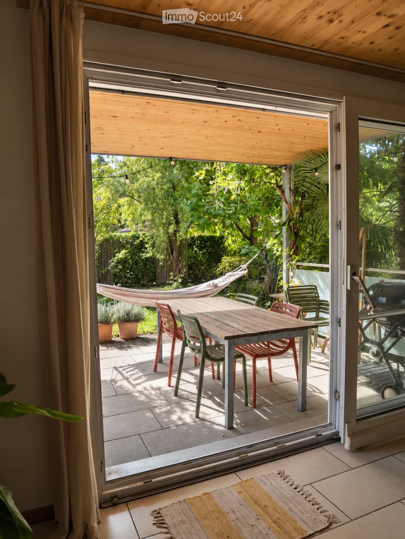 Open sliding glass door leads to a wooden deck with a dining table, chairs, and a hammock, surrounded by lush greenery.