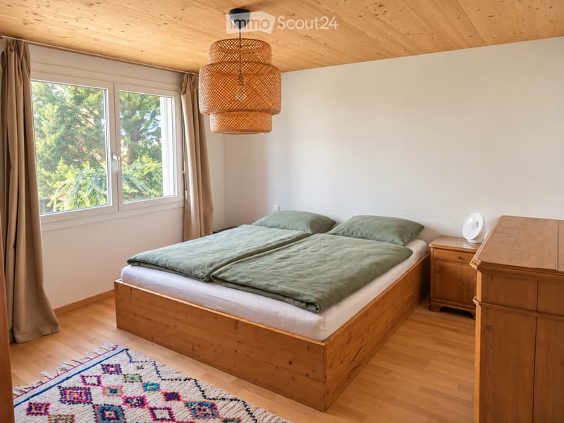 Cozy bedroom with wooden bed, green bedding, nightstand, and woven pendant light. Large windows offer a view of greenery outside.