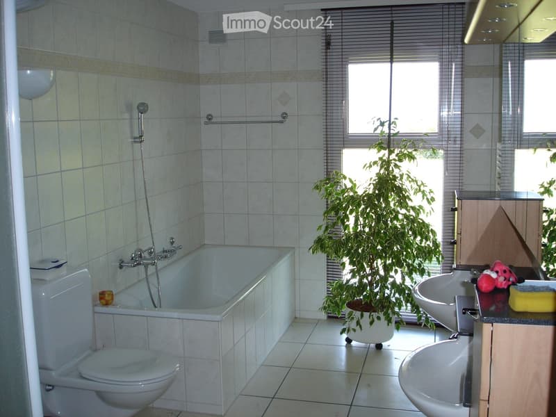 Modern bathroom with white tiles, bathtub, toilet, sink, and a potted plant near the window.