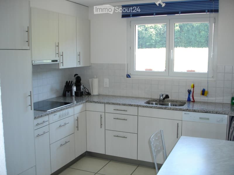 Modern kitchen with white cabinets, granite countertops, stainless steel sink, dishwasher, and a small dining area with a chair and table.