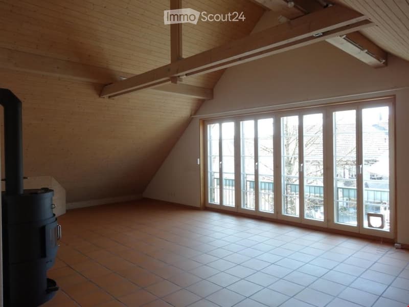 Empty room with wooden ceiling beams, large sliding glass doors leading to a balcony, tiled floor, and a wood-burning stove.