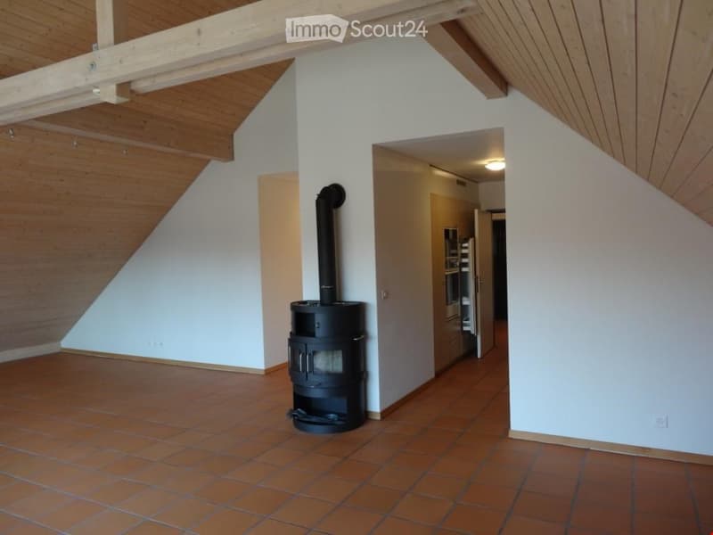 Spacious room with wooden ceiling beams, tiled floor, and a black wood-burning stove. The room has white walls and an open doorway leading to another area.