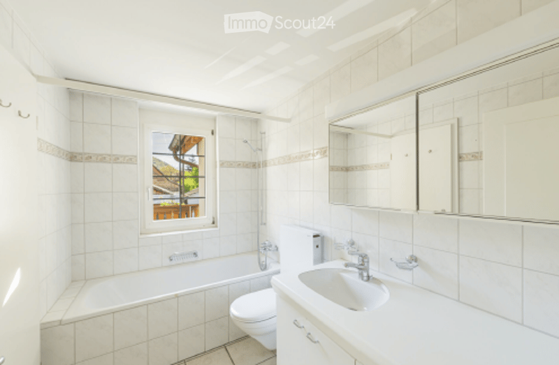 Bright bathroom with white tiles, a bathtub, toilet, sink, and a large mirror above the sink. A window offers a view of the outdoor area.