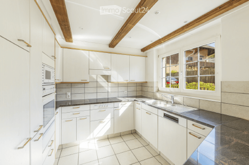 Bright, modern kitchen with white cabinets, stainless steel appliances, and exposed wooden beams on the ceiling. Features a large window with a view of the garden.