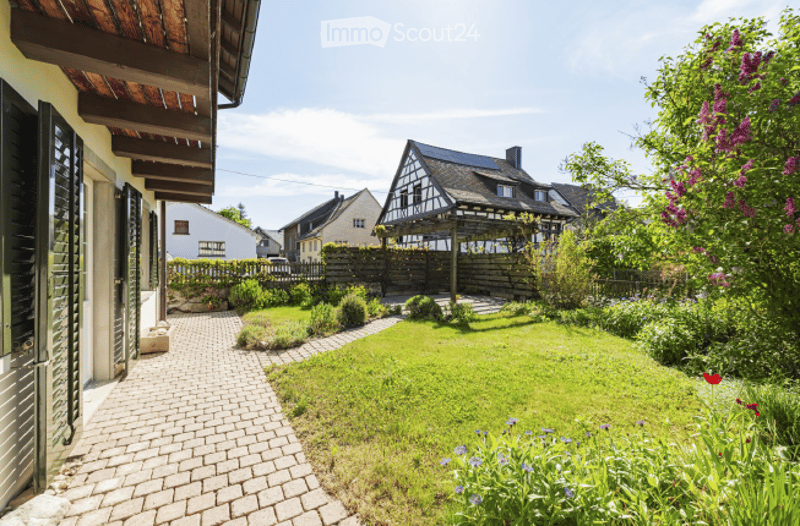 Well-maintained garden with a brick pathway leading to a wooden deck, surrounded by greenery and colorful flowers.
