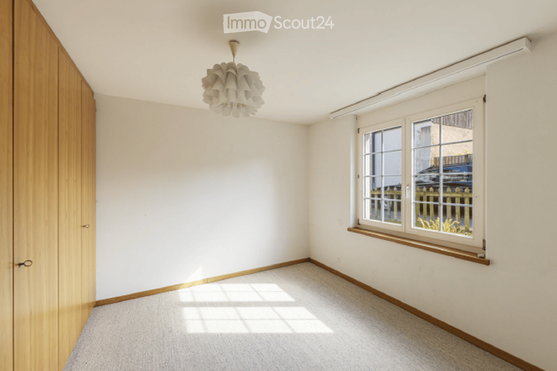 Empty room with white walls, wooden floor, large window with blinds, and a modern pendant light fixture.