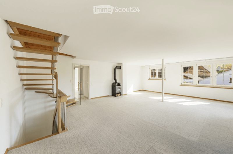 Empty room with white walls, wooden staircase, fireplace, and large windows.
