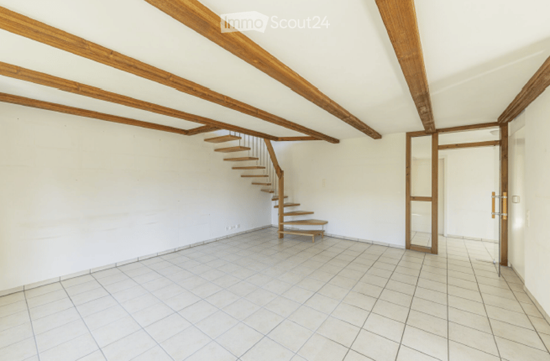 Empty room with white walls, exposed wooden beams, tiled floor, and a small wooden staircase leading to an upper level.