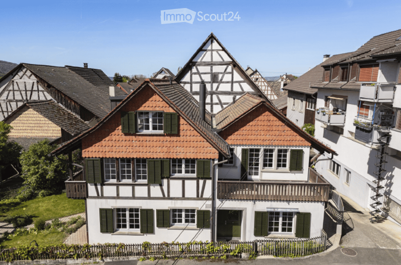 Traditional half-timbered house with red-tiled roof, green shutters, and white walls, surrounded by neighboring houses and greenery.
