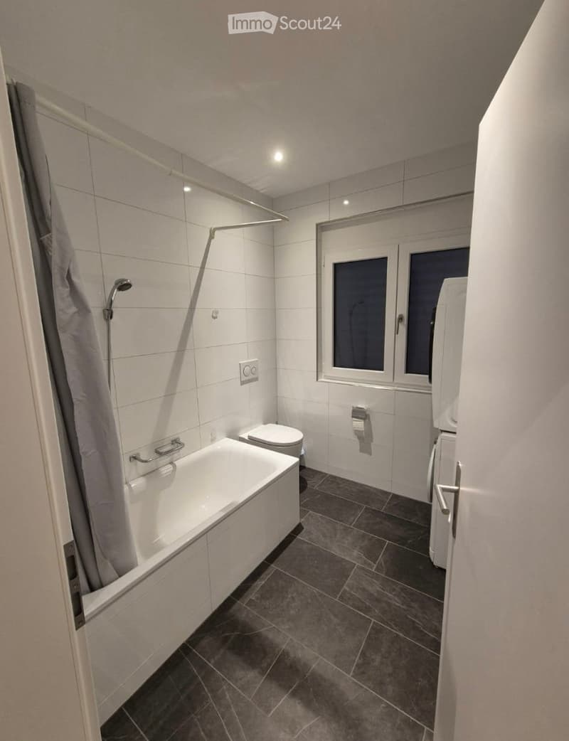Modern bathroom with a bathtub, toilet, shower, and a window with frosted glass. The room features white tiles and dark grey flooring.