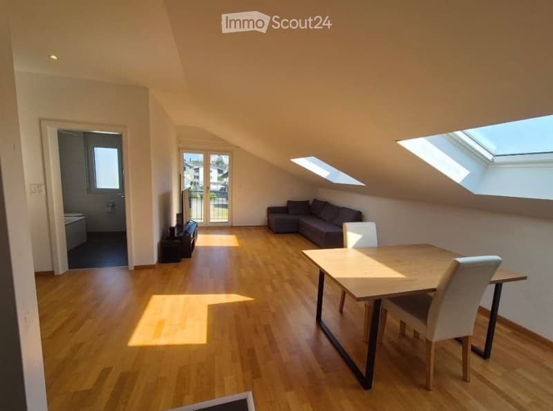 Bright, open-plan living and dining area with wooden flooring, large windows, skylights, and modern furniture including a sofa, dining table, and chairs.