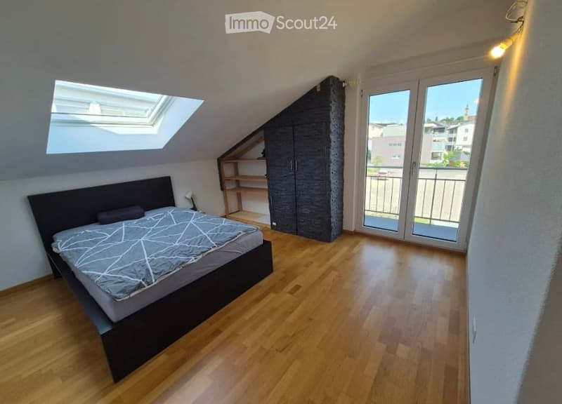 Spacious attic bedroom with wooden flooring, large window, skylight, and sliding glass doors leading to a balcony. Features a modern bed, wardrobe, and built-in shelves.