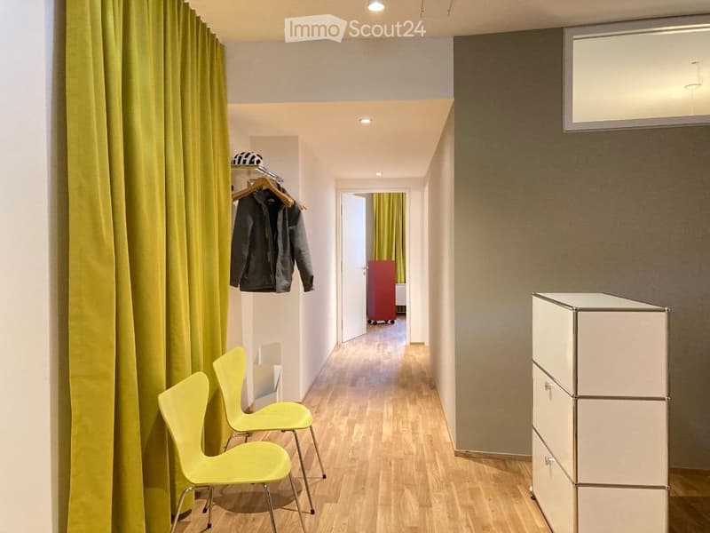 A narrow hallway with wooden flooring, two bright yellow chairs, a white cabinet, and a coat rack with jackets hanging on it. A doorway leads to another room with a red wardrobe and yellow curtains.