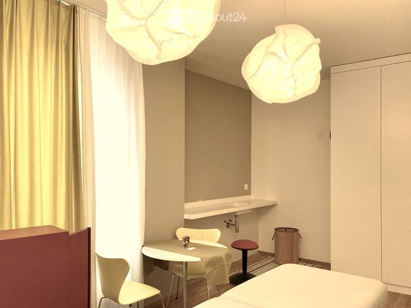 A modern, compact room with a bed, a small table with chairs, a sink area, and contemporary lighting fixtures.