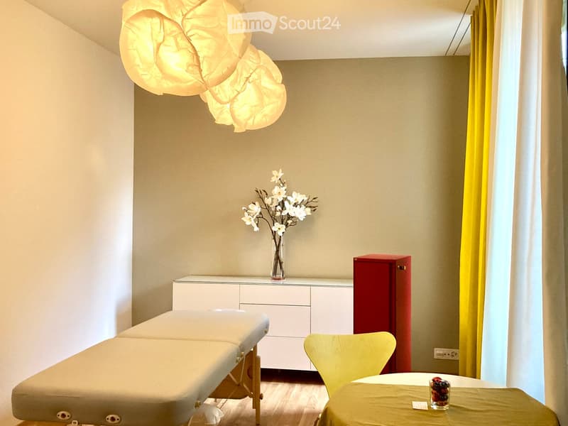 A well-lit room with a massage table, a yellow chair, a small table with a vase of white flowers, and a red cabinet.