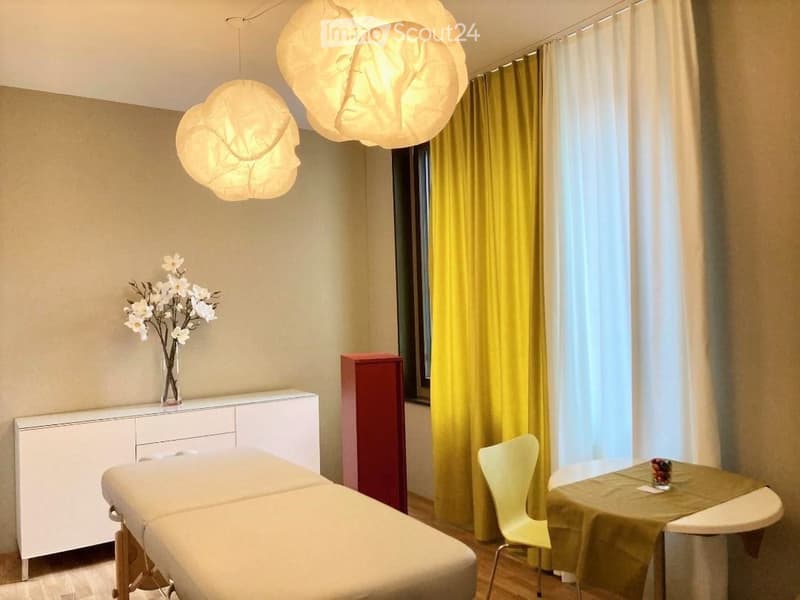 A modern, well-lit room with a massage table, a white cabinet, a small table with a green tablecloth, a yellow chair, and decorative flowers in a vase. The room features large windows with yellow curtains and unique hanging lamps.