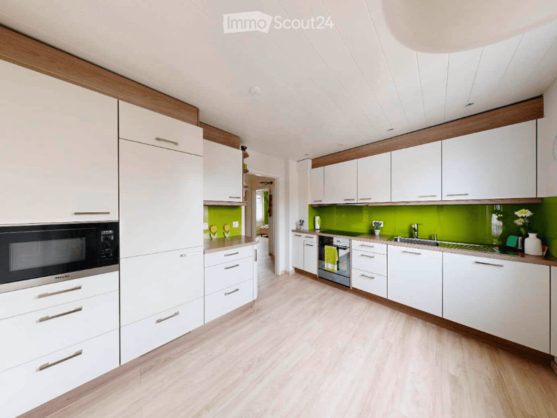 Modern kitchen with white cabinets, wooden accents, green backsplash, and stainless steel appliances including a microwave and oven. The floor is light wood, and the countertops are light-colored.