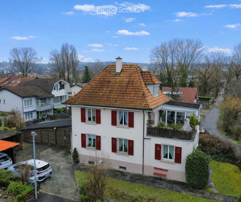 A two-story house with a red-tiled roof, white walls, and red shutters. The house features a balcony on the upper floor, a small garden area, and a driveway with parked cars.