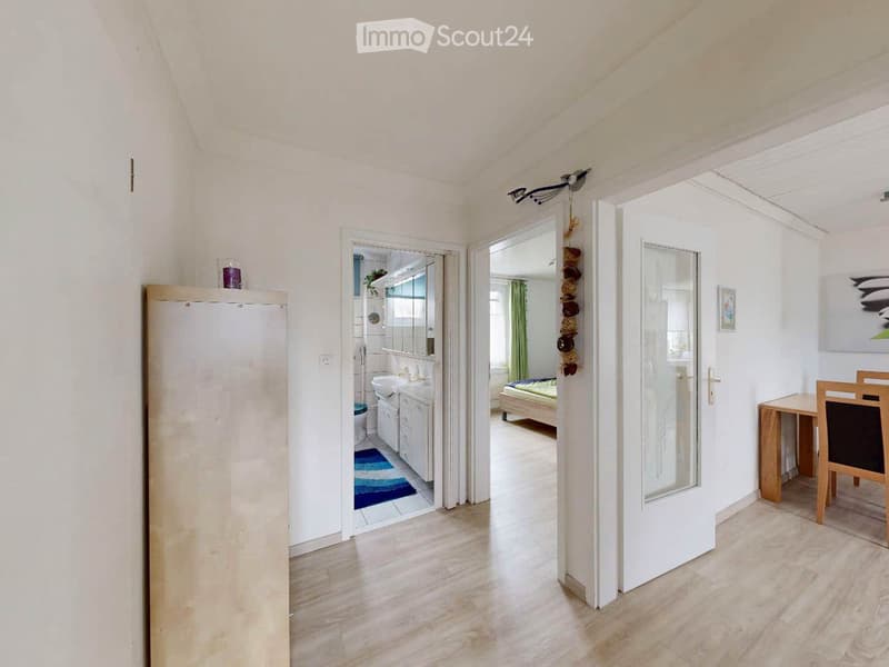 A well-lit hallway with light wooden flooring, white walls, and several doors leading to different rooms including a bathroom, bedroom, and dining area.