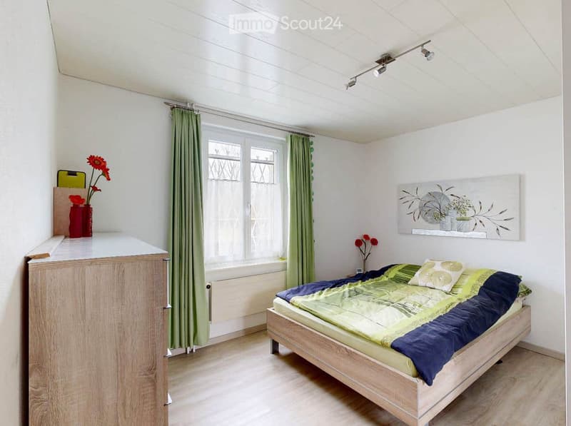 Bright bedroom with a wooden bed, green and blue bedding, a nightstand with a vase of red flowers, a painting on the wall, and green curtains on the window.