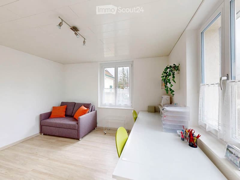 A modern, well-lit living room with a grey sofa, orange cushions, a white desk, and a green chair. The room features large windows, a plant, and a clock on the wall.