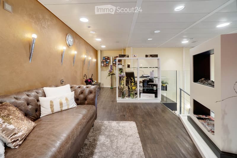 Modern waiting area with a brown leather sofa, decorative pillows, a small table with a vase of flowers, a glass display case, and a sleek office area with a chair and computer.