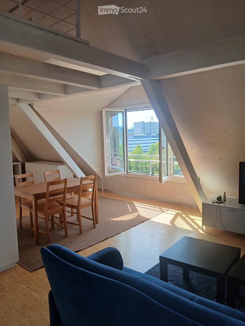 Bright, open living and dining area with wooden floors, large windows, and skylights. Features a blue sofa, coffee table, dining table with chairs, and a small cabinet.