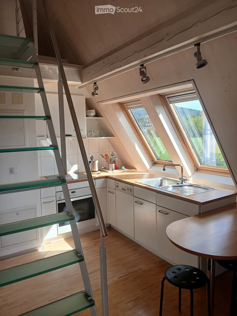 Modern kitchen with white cabinets, wooden floor, and large windows. Features a sink, oven, dishwasher, and a small dining area with a table and stool.