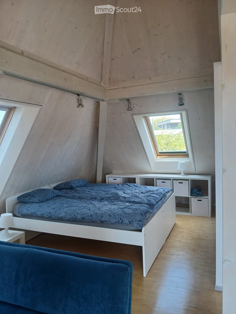 Cozy attic bedroom with a white bed, blue bedding, a blue sofa, a white nightstand with a lamp, and a white storage unit with multiple compartments.