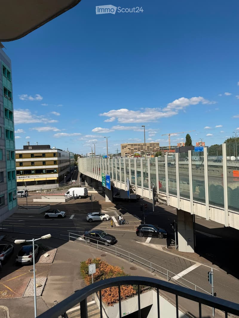 View from a balcony overlooking a parking lot with several cars, a motorcycle, and a few buildings in the background under a clear blue sky.