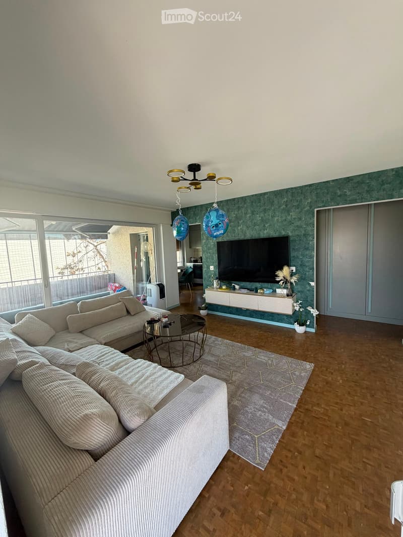 Modern living room with a large sectional sofa, coffee table, television, and decorative vases. The room features wooden flooring, a grey rug, and large windows with a view of the balcony.