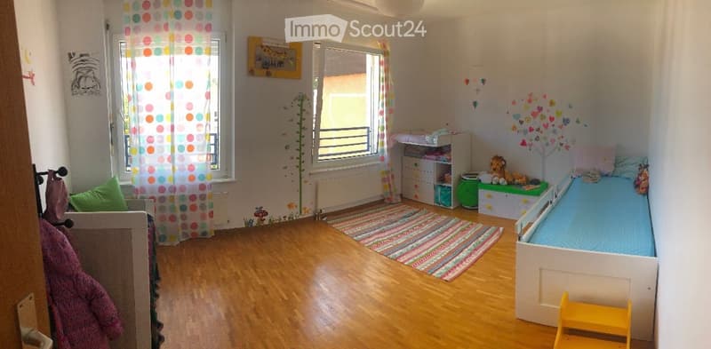 Child's bedroom with a single bed, colorful rug, storage unit, and various toys. The room features wooden flooring, large windows with colorful curtains, and wall decorations including a tree and heart shapes.
