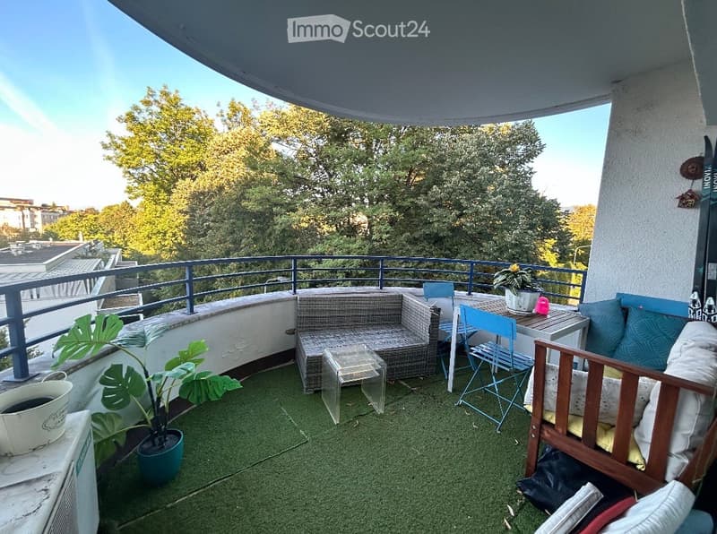 Balcony with wicker sofa, small table, chairs, potted plants, and a view of trees and buildings.