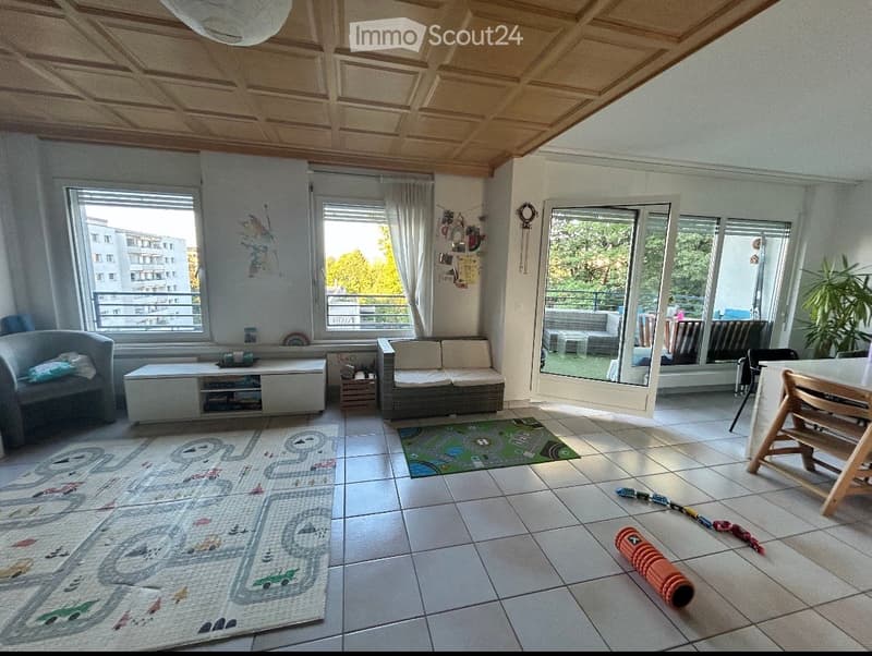 Bright living room with large windows and a sliding glass door leading to a balcony. Features a sofa, armchair, coffee table, and a small dining area with a table and chairs. The floor is tiled, and there are children's play mats and toys on the floor.