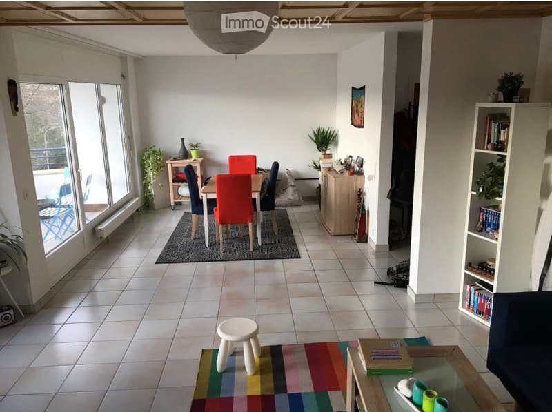 Open-plan living and dining area with tiled flooring, large windows, colorful rugs, and modern furniture including a dining table, chairs, bookshelf, and a small stool.