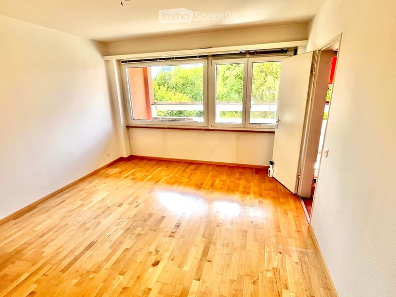 Empty room with wooden flooring, white walls, large windows, and an open doorway leading to another space.
