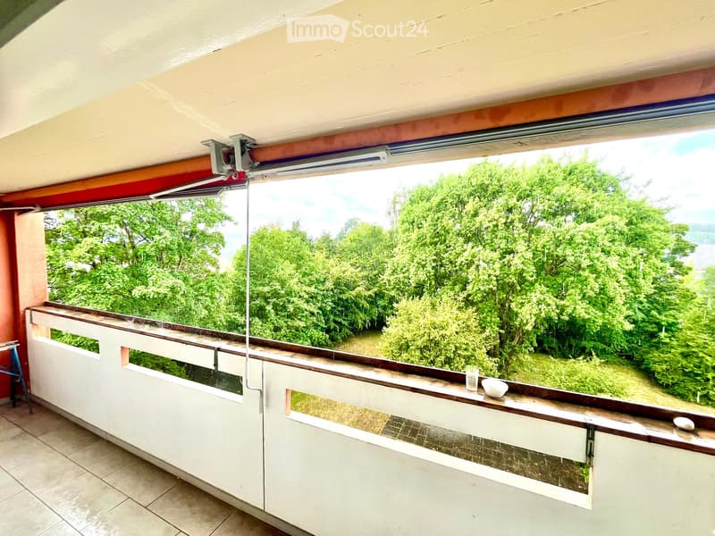 Balcony with wooden railing, tiled floor, and a scenic view of green trees and a body of water.