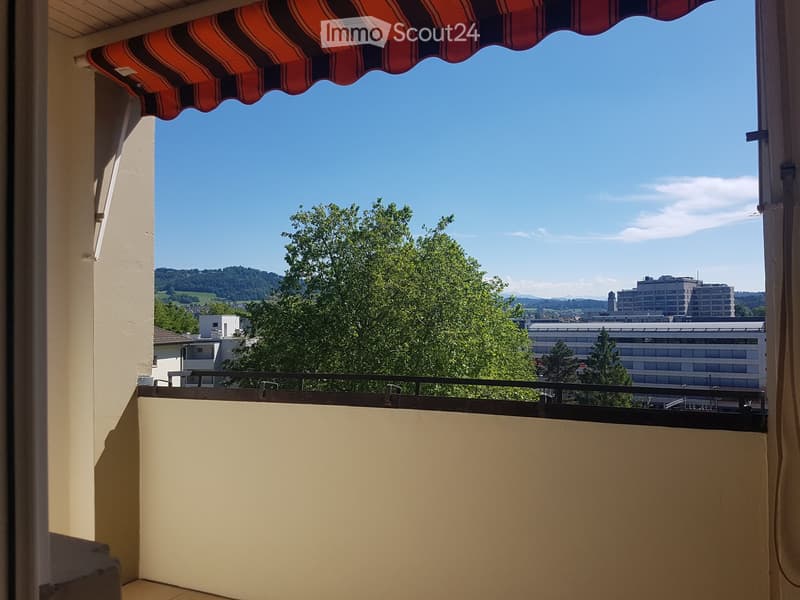 Balcony with striped awning, metal railing, and scenic view of trees, buildings, and distant hills.